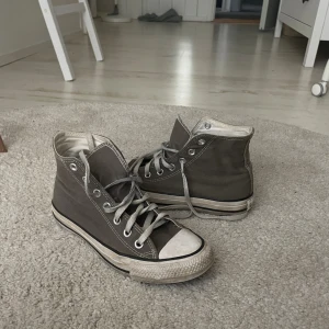 Grå Converse Chuck Taylor All Star - Klassiska Converse Chuck Taylor All Star i grått canvasmaterial med vita och svarta detaljer. Högt skaft, rund tå och snörning framtill. Ikonisk logga på sidan och vit sula med svart rand. Perfekta sneakers för en avslappnad och cool stil.