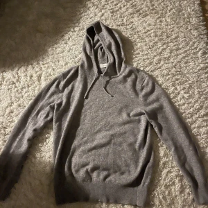 Grå cashmere hoodie från Sayless - Mjuk och lyxig grå hoodie från Sayless i 100% cashmere. Tröjan har huva med snörning och långa ärmar, perfekt för dig som gillar stilren och chill look. Ribbstickade muddar vid ärmslut och nederkant ger extra komfort. Snygg att matcha med jeans eller joggers.