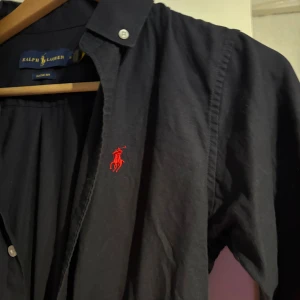 Mörkblå skjorta från Ralph Lauren M - Klassisk mörkblå skjorta från Ralph Lauren med röd broderad logga på bröstet. Skjortan har button-down krage, vita knappar och är tillverkad i bomull. Perfekt för dig som gillar stilrena och tidlösa plagg.