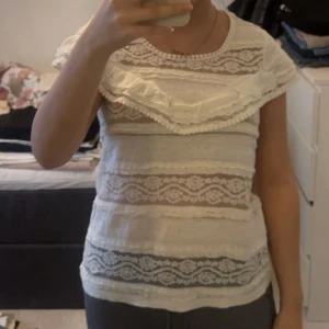 Vit spetsblus med volang från Lindex - Säljer en söt vit blus från Lindex med spetsdetaljer och volang runt axlarna. Blusen har rund halsringning och är ärmlös, med genombrutna spetsmönster i horisontella ränder. Perfekt för dig som gillar romantisk och feminin stil.