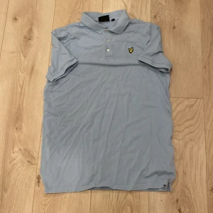 Ljusblå piké från Lyle & Scott - Fräsch ljusblå pikétröja från Lyle & Scott med klassisk krage och två knappar framtill. På bröstet sitter den ikoniska broderade gula örnen. Tröjan har korta ärmar och är gjord i mjuk bomull som är skön mot huden. Perfekt för dig som gillar stilren och enkel design.