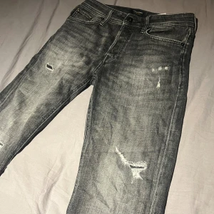 Svarta jeans med slitningar, storlek 29/32 - Snygga svarta jeans från Jack & Jones i storlek 29/32. Jeansen har coola slitningar och en tvättad look som ger dem en rå vibe. Klassisk femficksmodell med knapp och dragkedja. Perfekta för dig som gillar streetstyle.