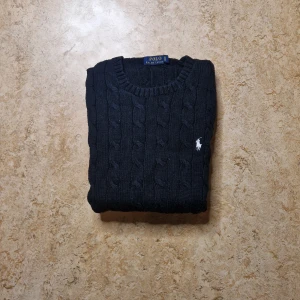 Svart stickad tröja Polo Ralph Lauren - Svart kabelstickad tröja från Polo Ralph Lauren med klassisk rund hals och vit broderad logga på bröstet. Tröjan har ribbade muddar vid ärmslut och nederkant, och är tillverkad i mjukt stickat material. Perfekt för dig som gillar stilren och tidlös design.