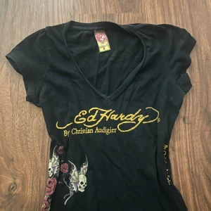 Svart Ed Hardy topp med v-ringning - Svart topp från Ed Hardy med v-ringad hals och korta ärmar. Framsidan har ett stort gult Ed Hardy-tryck och coola detaljer med döskallar, rosor och vingar i lila, rött och beige. Materialet är mjukt och stretchigt, perfekt för dig som gillar edgy streetstyle.