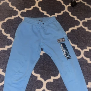 Blå Hoodrich mjukisbyxor med snörning - Snygga ljusblå mjukisbyxor från Hoodrich med tryckt logga och patch på ena benet. Byxorna har snörning i midjan och praktiska fickor, varav en med dragkedja. Perfekta för chill eller streetwear-stil.