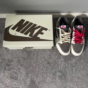 Nike Air Jordan 1 Low Travis Scott - Säljer ett par Nike Air Jordan 1 Low Travis Scott med beige och bruna paneler, ikonisk omvänd Swoosh och röda broderade detaljer på hälen. Skorna har platt sula och rund tå, samt levereras med både beige och röda skosnören.