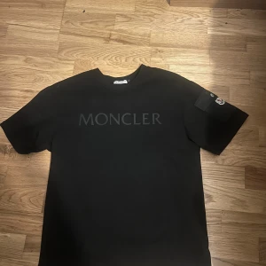 Svart Moncler t-shirt med ficka - Svart t-shirt från Moncler med diskret logga på bröstet och en snygg ficka med Moncler-märke på ena ärmen. Klassisk passform och rund halsringning. Tillverkad i mjuk bomull som känns skön mot huden.