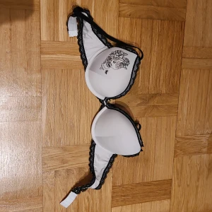 Vit BH med döskalle från Censored - Säljer en vit BH från Censored med svart spetskant och coolt döskalletryck med blommor och paljetter på ena kupan. Axelbanden är svarta och toppen har en snygg kontrast mellan vitt och svart. Perfekt för dig som gillar edgy detaljer!