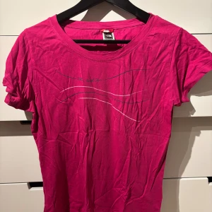 Rosa t-shirt från The North Face - Snygg rosa t-shirt från The North Face med tryck på bröstet och logga på ryggen. Modellen är klassisk med rund hals och korta ärmar. Tillverkad i mjuk bomull som känns skön mot huden. Perfekt för dig som gillar färg och vill sticka ut lite extra.