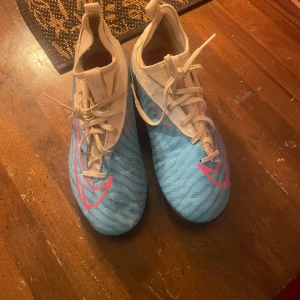 Blå fotbollsskor med snörning - Säljer ett par snygga fotbollsskor i ljusblått med mönstrade detaljer och rosa logga på sidan. Skorna har snörning och platt sula med dobbar för bra grepp på planen. Ovandelen är i syntetmaterial och insidan är vit. Perfekt för dig som vill sticka ut på matchen!