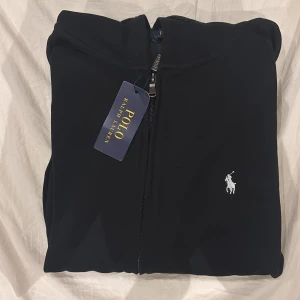 Svart hoodie från Polo Ralph Lauren - Svart hoodie från Polo Ralph Lauren med klassisk vit broderad logga på bröstet. Tröjan har huva med snörning, dragkedja framtill och stora fickor. Tillverkad i mjuk bomull som är skön att bära. Perfekt för dig som gillar stilrena och tidlösa plagg. Helt ny med prislapp kvar och allt.