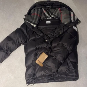 Svart pufferjacka från Burberry - Svart pufferjacka från Burberry med klassisk logga på ärmen och rutigt foder i huvan. Jackan har dragkedja och knappar framtill, samt snörning nedtill. Materialet är polyamid med dunfyllning för extra värme. Perfekt för kalla dagar och har en riktigt snygg streetstil.