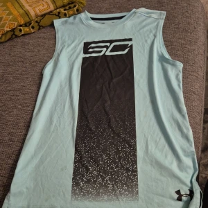 Under Armour SC ärmlös träningströja - Ljusblå ärmlös träningströja från Under Armour med stort svart SC-tryck framtill och HeatGear-material som håller dig sval. Loose fit för skön rörelsefrihet, perfekt för gymmet eller sport. Under Armour-logga nere till höger.