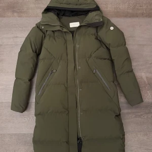 Olivgrön lång dunjacka Sail Racing - Snygg olivgrön lång dunjacka från Sail Racing med huva och två snedställda dragkedjefickor. Jackan har quiltad design, logotyp på ärmen och robust dragkedja framtill. Perfekt för kalla dagar med stilren look och skön passform.