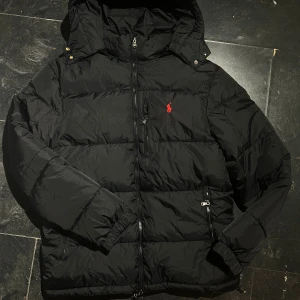 Svart pufferjacka från Polo Ralph Lauren - Svart pufferjacka från Polo Ralph Lauren med röd broderad logga på bröstet. Jackan har huva, flera dragkedjor och praktiska fickor. Materialet är syntet och jackan är quiltad för extra värme. Perfekt för kalla dagar och har en clean, modern look.