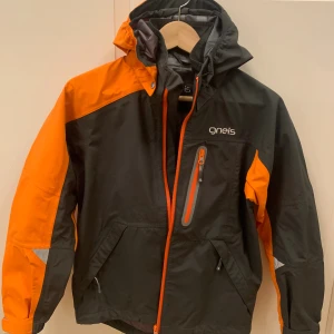 Svart/ orange regn- och vindjacka från Gneis - Säljer en Gneis Abisko Jacket i svart och orange med huva och flera fickor. Jackan har dragkedja framtill och coola detaljer som reflekterande partier. Materialet är 100% polyamid, perfekt för utomhusaktiviteter. Märket syns både fram och bak på jackan.