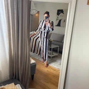 Randig omlottklänning från Lindex - Säljer en lång omlottklänning från Lindex med snygga vertikala ränder i marinblått, vitt och rött. Klänningen har trekvartsärm, v-ringning och knytband i midjan. Perfekt för dig som gillar en chill och trendig look med lite extra edge.