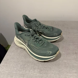 Gröna Hoka One One sneakers 42,5 - Säljer ett par gröna Hoka One One sneakers med tjock, vit sula och snygg logga på sidan. Ovandelen är i mesh för bra andningsförmåga och skorna har snörning. Perfekta för dig som gillar streetstyle och komfort.