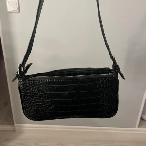 Svart axelväska med krokoprägling - Snygg svart axelväska med krokopräglat mönster och justerbar rem med spänne. Väskan har en praktisk dragkedja upptill. Perfekt storlek för det viktigaste och passar till många olika stilar.