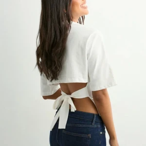 Vit topp från nelly - Säljer en vit croppad topp med korta ärmar och rund hals. Toppen har en öppen rygg med breda band som knyts i en rosett baktill, vilket ger en snygg och trendig look. Materialet känns som mjuk bomull och passformen är normal. Tröjan är aldrig använd