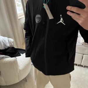 Svart PSG x Jordan vindjacka M - Den är i storlek L men sitter mer som en M, Svart vindjacka från Jordan x PSG med huva, dragkedja och snygga loggor på bröstet. Jackan har snörning vid huvan och coolt mönstrat innerfoder. Perfekt för dig som gillar sportig streetstyle och vill sticka ut med en fet collab.