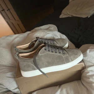 Grå mocka sneakers Christian Louboutin - Snygga grå sneakers från Christian Louboutin i mjuk mocka med klassisk vit platt sula och ikonisk röd undersida med logotyp. Skorna har rund tå och grå snörning, perfekt för dig som gillar exklusiv streetstyle och vill sticka ut med detaljer.