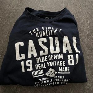 Mörkblå sweatshirt från Smog - Mörkblå sweatshirt från Smog med stort vitt tryck framtill där det står 'CASUAL' och flera vintage-inspirerade texter. Tröjan har rund halsringning och långa ärmar. Perfekt för en avslappnad streetstyle-look.