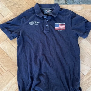 Mörkblå Polo Club pikétröja S - Snygg mörkblå pikétröja från Polo Club med klassisk krage och knappar. Tryck med amerikansk flagga och texten 'Royal Polo Saint Vincent' på bröstet. Perfekt för dig som gillar sportig och stilren look. Materialet är mjuk bomull som känns skönt mot huden.