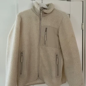 Beige teddyjacka från A Day's March - Säljer en beige teddyjacka från A Day's March i storlek Small. Jackan har mjukt fleece-material, hög krage och tre praktiska fickor med dragkedja framtill. Perfekt för dig som gillar en chill och cozy stil under höst och vinter.