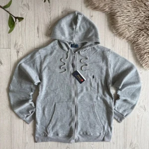 Grå hoodie från Polo Ralph Lauren - Snygg grå hoodie från Polo Ralph Lauren med dragkedja och huva. Tröjan har långa ärmar, två fickor fram och klassisk Polo-logga broderad på bröstet. Perfekt för chill dagar och enkel att matcha med jeans eller joggers.