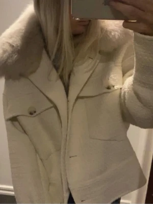Vit med stor pälskrage - Säljer min vita Meotine jacka med beige päls. Jackan har två stora bröstfickor med knappar och en rak, boxig passform. Materialet är strukturerat och ger en lyxig känsla. Perfekt för dig som vill sticka ut med en trendig och mysig jacka. Jackan är väldigt fint skick men längst ner på ärmen som saknas det en knapp. Det syns inte alls. Bilderna är inte mina egna men skickar gärna egna!
