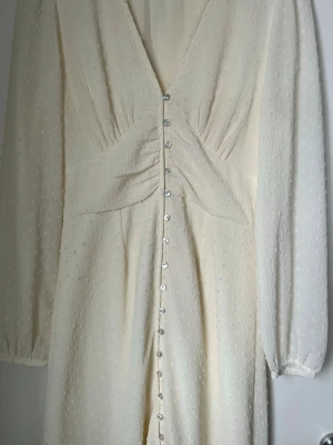Vit långklänning med knappar från Gina Tricot - Supersöt långklänning i vit ton från Gina Tricot. Klänningen har v-ringning, långa ärmar med resår vid ärmslut och fina knappar hela vägen framtill. Materialet är lätt strukturerat med diskreta prickar för extra detalj. Perfekt för sommarens alla tillfällen!