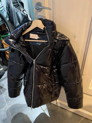 Svart pufferjacka från Nelly - Säljer en svart, glansig pufferjacka från Nelly i storlek S. Jackan har hög krage, dragkedja framtill och stora fickor. Insidan är fodrad med mjukt material för extra värme. Perfekt för kalla dagar och riktigt snygg till streetwear.