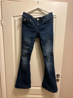 True Religion blå bootcut jeans - Säljer ett par ikoniska blå jeans från True Religion med bootcut passform och låg midja. Jeansen har snygga kontrastsömmar och guldiga knappar på fickorna bak, samt det klassiska broderade mönstret på bakfickorna. Tillverkade i slitstarkt denim. Storlek 27/32