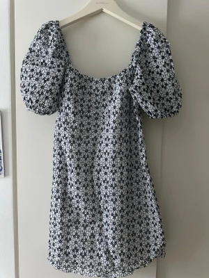 Blommig kortklänning från & Other Stories - Supersöt kortklänning från & Other Stories med puffärmar och broderat blommönster i svart på vit botten. Klänningen har en rundad urringning och är tillverkad i bomull, perfekt för en trendig och fräsch look.