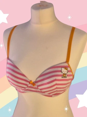 Hello Kitty rosarandig BH 75B - Söt behå med rosa och vita ränder från Hello Kitty. Behån har tunna orange axelband och en liten rosett mellan kuporna och ett tryck av Hello Kitty på ena sidan. Perfekt för en lekfull och färgglad stil. Varan är oanvänd och kommer från ett konkurslager från 2000-talet. Har fler annonser och bra bundlerabatt vid köp av flera.