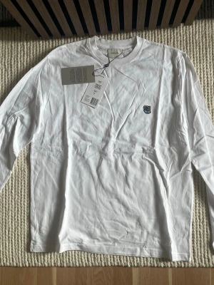 Vit långärmad tröja Tonsure Teddy - Säljer en clean vit långärmad tröja från Tonsure med färgglatt teddy-logomärke på bröstet. Tröjan är i mjuk bomull och har en relaxed fit med rund hals. Perfekt för dig som gillar minimalistisk stil med en twist. Finns i olika storlek 2st L och 1st XL