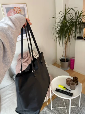 Svart axelväska i skinn - Säljer en stilren svart axelväska med långa handtag och vågig kant upptill. Väskan är rymlig och passar perfekt för dig som vill ha plats med allt du behöver. Tillverkad i skinn och har en enkel, klassisk design utan synliga loggor.