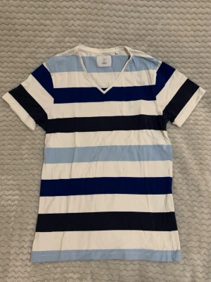 Randig t-shirt från Bläck - Snygg t-shirt från Bläck med breda horisontella ränder i vitt, marinblått och ljusblått. T-shirten har v-ringning och korta ärmar, perfekt för en avslappnad stil. Tillverkad i mjuk bomull som känns skön mot huden.