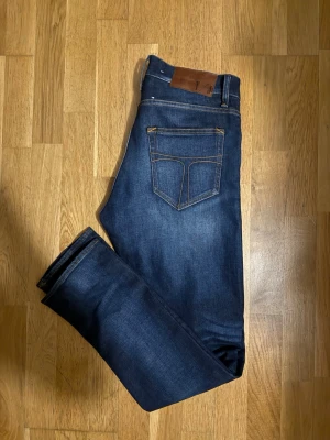 Mörkblå washed jeans från Tiger of Sweden - Säljer ett par mörkblå jeans från Tiger of Sweden med klassisk femficksdesign och snygga kontrastsömmar. Jeansen har rak passform och är tillverkade i slitstarkt bomullsmaterial. Perfekta för dig som gillar stilrena och tidlösa jeans.