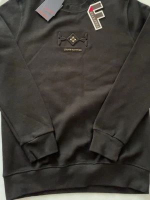 Svart Louis Vuitton sweatshirt - Säljer en svart sweatshirt från Louis Vuitton med broderad logga och diskret mönster på bröstet. Tröjan har rund halsringning och långa ärmar. Materialet känns mjukt och är troligtvis bomull. Perfekt för dig som gillar exklusiva streetwear-plagg.