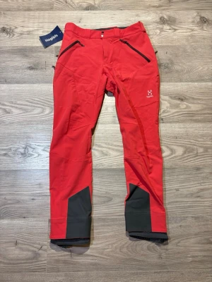 Haglöfs  - Snygga röda täckbyxor från Haglöfs med svarta förstärkningar vid bensluten och dragkedjeförsedda fickor framtill. Byxorna har normal passform och är tillverkade i slitstarkt syntetmaterial, perfekta för vinteraktiviteter.