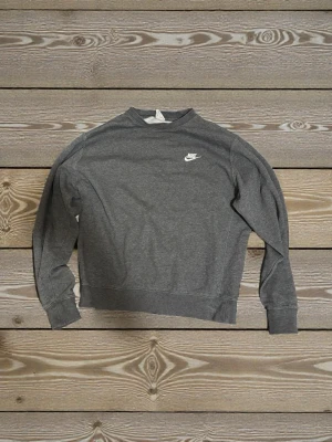 Mörkgrå sweatshirt från Nike - Mörkgrå sweatshirt från Nike med klassisk logga broderad i vitt på bröstet. Tröjan har rund hals, långa ärmar och ribbade muddar vid ärmslut och nederkant. Perfekt för chill dagar eller när du vill ha en clean look.