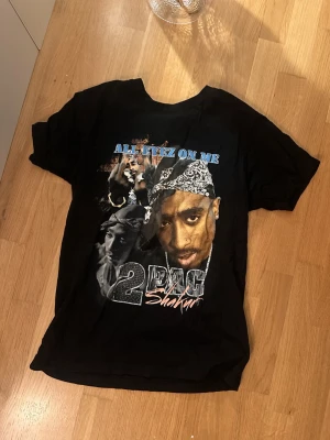 Svart 2Pac t-shirt All Eyez On Me - Svart t-shirt med tryck av 2Pac och texten 'All Eyez On Me' på framsidan. Klassisk passform med korta ärmar och rund hals. Materialet känns som bomull och trycket har en cool vintage-vibe. Perfekt för dig som gillar hiphop och streetstyle.