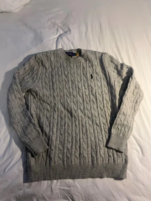 Grå kabelstickad tröja från Polo Ralph Lauren - Säljer en grå kabelstickad tröja från Polo Ralph Lauren med klassisk svart logga på bröstet. Tröjan har rund halsringning, långa ärmar och ribbade muddar. Perfekt för dig som vill ha en stilren och tidlös look. Sitter som en XL