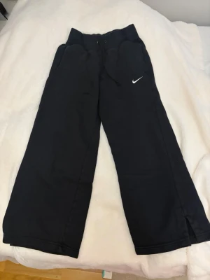 Svarta mjukisbyxor från Nike S - Svarta mjukisbyxor från Nike med bred resår i midjan och snörning. Byxorna har en relaxed passform med raka, vida ben och liten slits vid benslutet. Klassisk Nike-logga broderad i vitt på ena benet. Perfekta för chill dagar eller träning. (allting tvättas innan leveran)