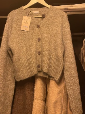 Grå croppad kofta från Zara  - Mysig grå kofta från zara i croppad modell. Tröjan har långa ärmar, ribbade muddar och stora knappar framtill. Perfekt för lager på lager och ger en chill vibe till din outfit.