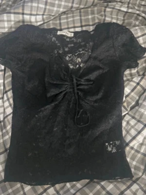 Svart spetsblus med snörning Wild Love - Säljer en svart blus från Wild Love i storlek M. Toppen är helt i spets med blommigt mönster, har korta ärmar och en snygg snörning framtill som ger en v-ringad look. Materialet är genomskinligt och stretchigt, perfekt för dig som gillar detaljer och trendig stil.