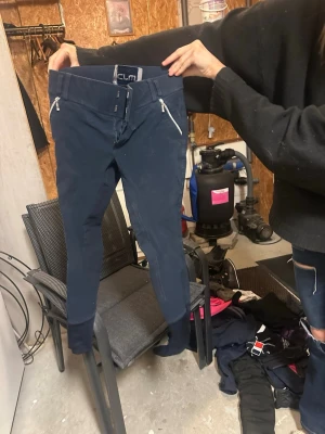 Mörkblå ridbyxor från CRW stl 150 - Säljer ett par mörkblå ridbyxor från CRW med dragkedjor framtill och förstärkta knän. Byxorna har smal passform och bred linning med hällor för bälte. Perfekta för ridning och snygga detaljer med silverfärgade dragkedjor.