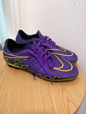 Nike Hypervenom lila fotbollsskor - Säljer ett par Nike Hypervenom fotbollsskor i lila med gula detaljer och svart sula. Skorna har snörning, mönstrad ovandel och dobbar för bra grepp på planen. Tillverkade i syntetmaterial och designade för att sticka ut på fotbollsplanen.